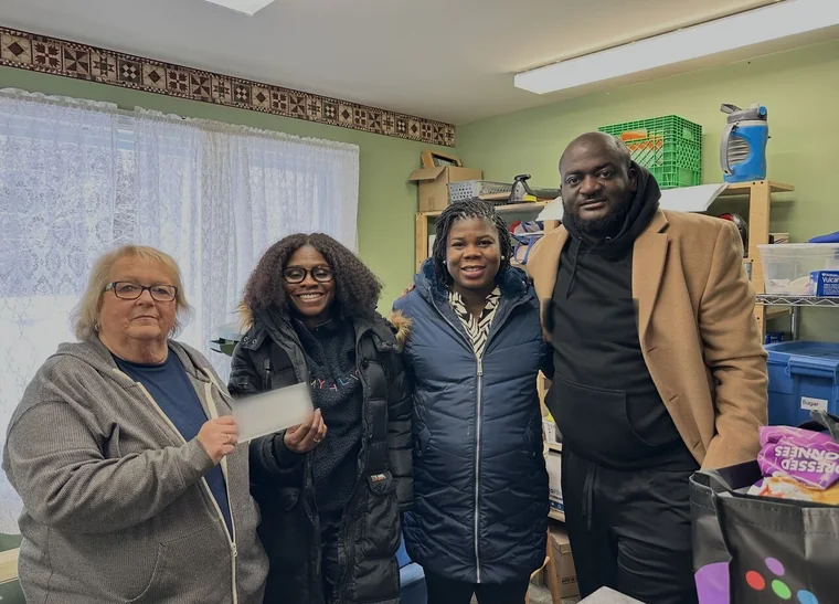 Four people stand together in the food bank's storage room. One holds the donation cheque.